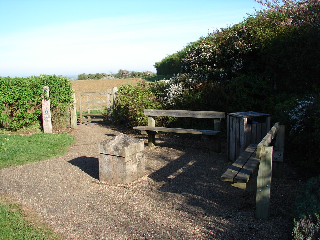 Two wooden benches at a rest stop on the Garden City Greenway, with a waymarker post, a field gate and farmland beyond